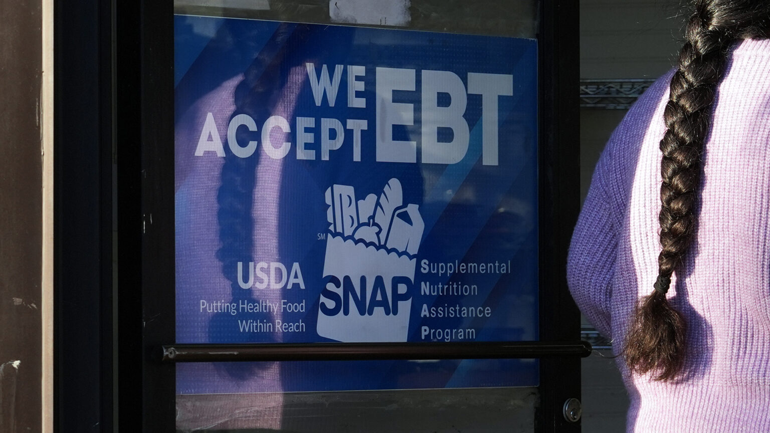 A woman walks through an open glass and metal door with a sign with a stylized logo of a grocery bag filled with an egg carton, apple, loaf of bread and milk carton along with the words We Accept EBT, USDA, Putting Healthy Food Within Reach and Supplemental Nutrition Assistance Program.