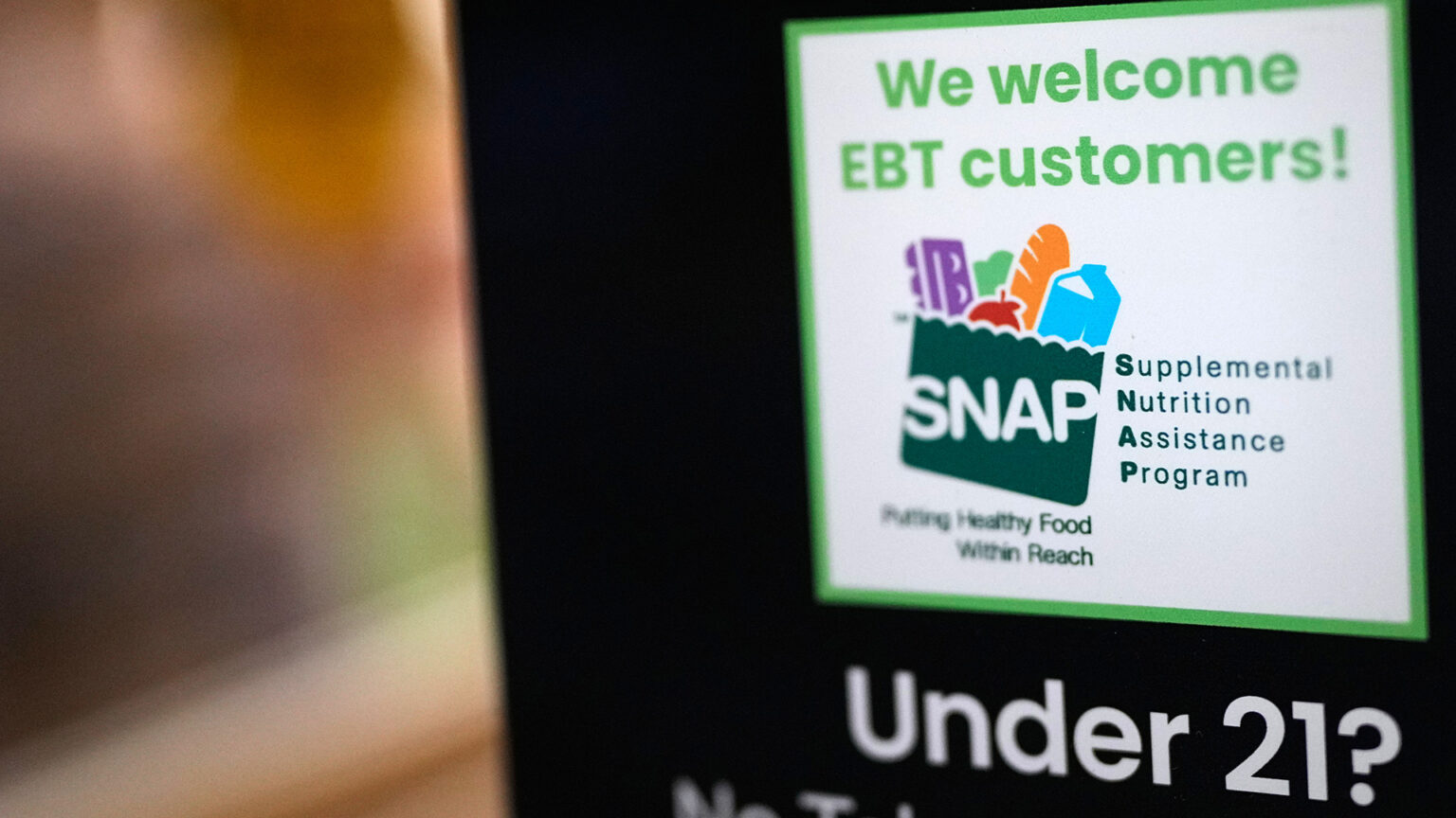 A sign with a stylized logo of a grocery bag filled with an egg carton, apple, loaf of bread and milk carton along with the words We welcome EBT customers!, Supplemental Nutritional Assistance Program, Putting Healthy Food Within Reach and Under 21? is displayed in a window.