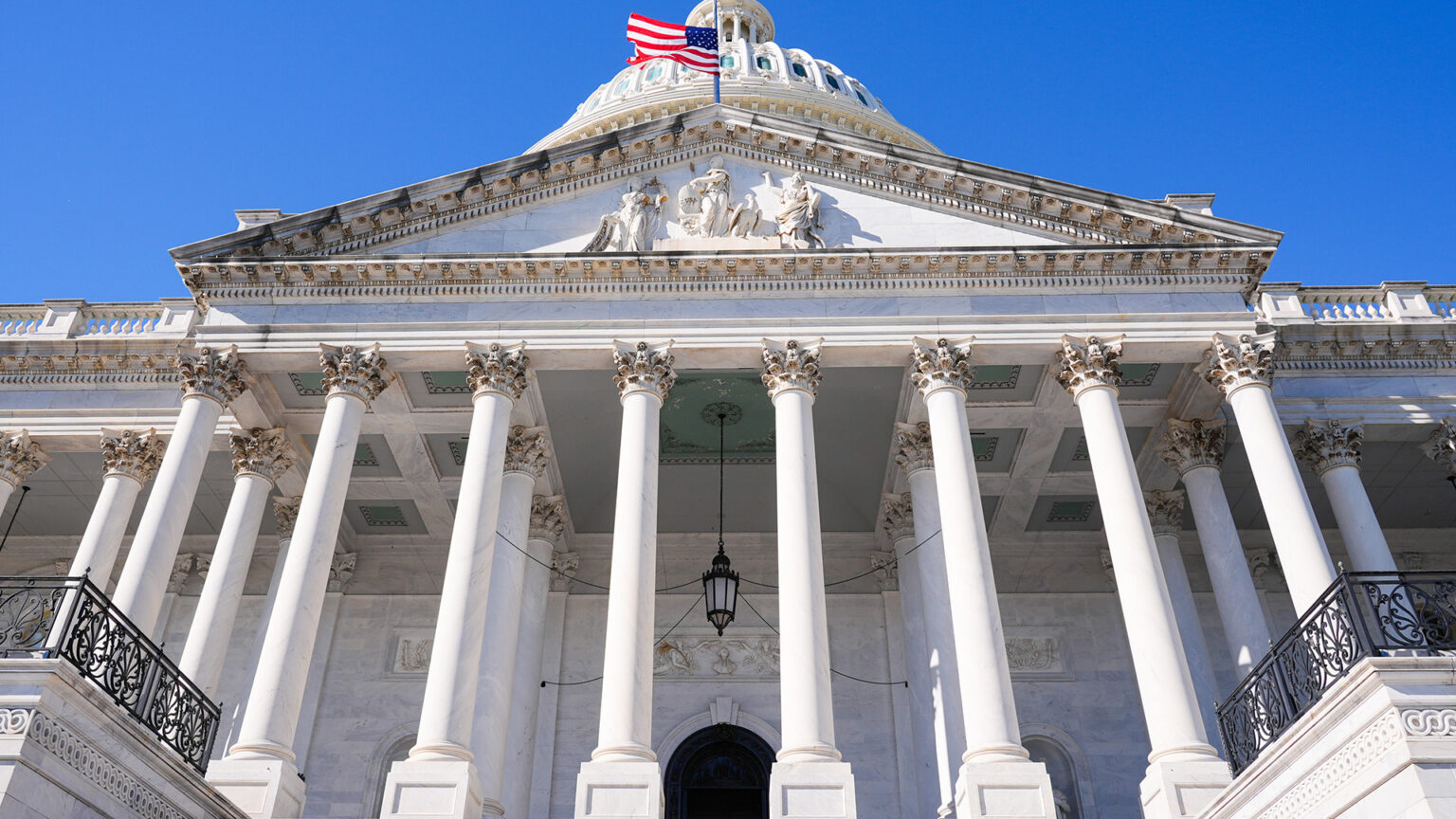 A flag at half staff flies on a flagpole above a pediment with multiple statues supported by multiple rows of columns of a Neoclassical masonry building with a central dome.