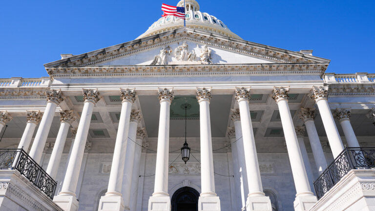 A flag at half staff flies on a flagpole above a pediment with multiple statues supported by multiple rows of columns of a Neoclassical masonry building with a central dome.