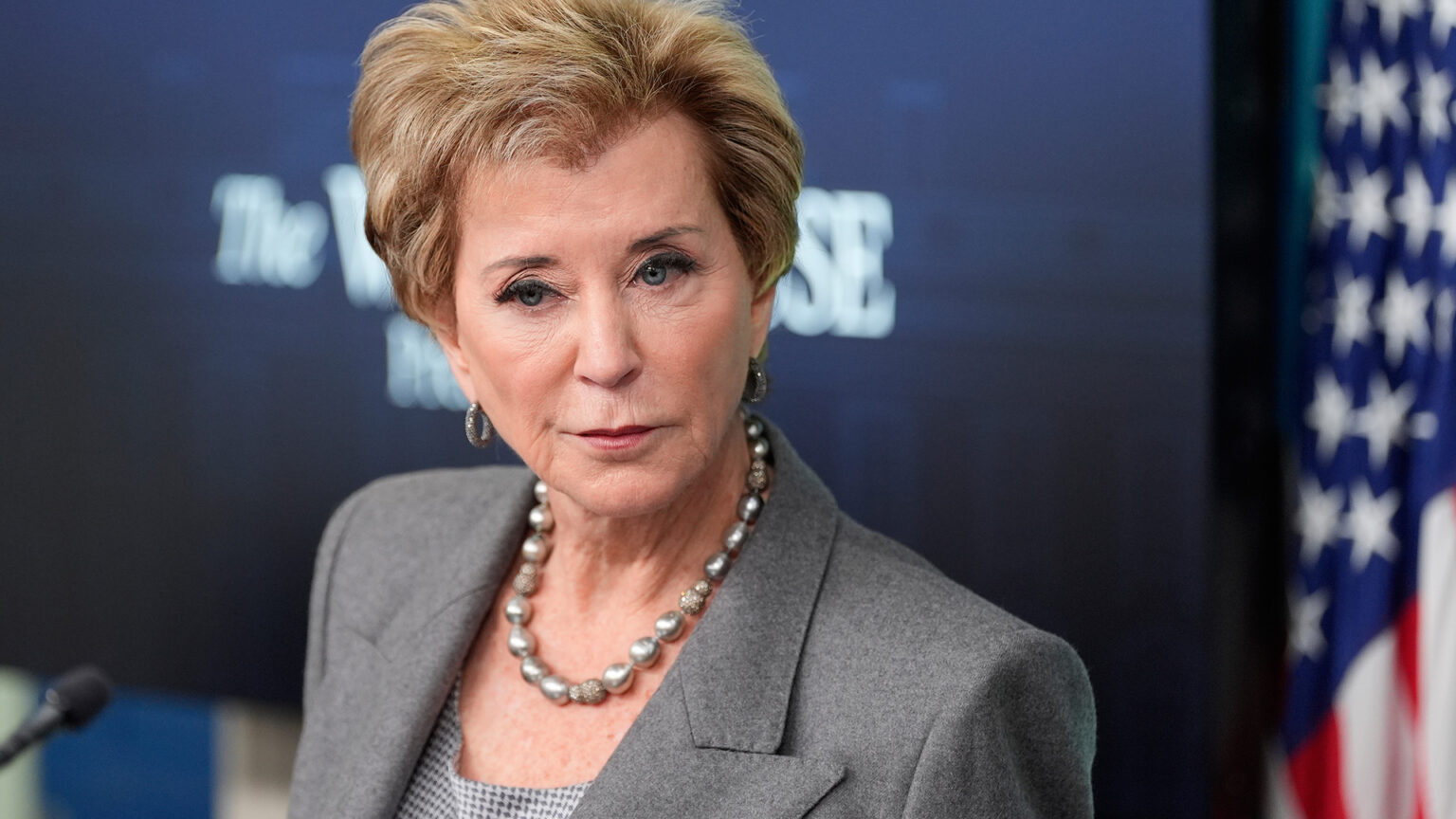 Linda McMahon speaks while standing in a room with an out-of-focus projector screen and U.S. flag in the background.