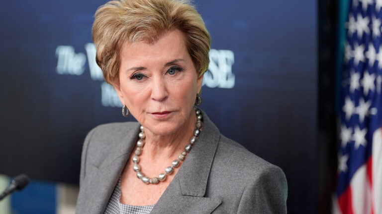 Linda McMahon speaks while standing in a room with an out-of-focus projector screen and U.S. flag in the background.