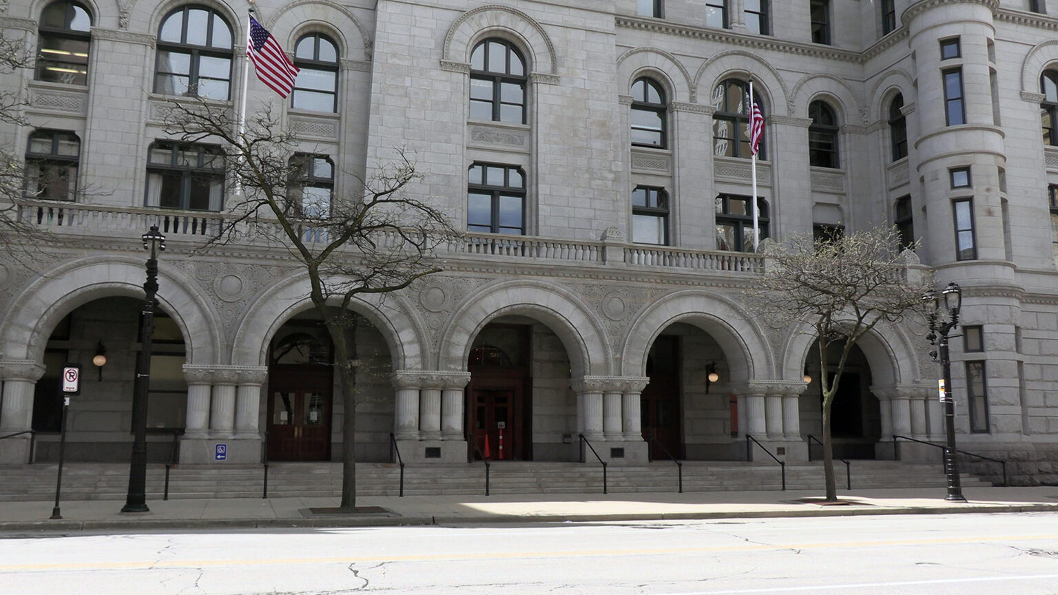 Two U.S. flags on flagpoles wave over a balustrade above a series of arches framing multiple doorways and windows on the ground floor of a multi-story Richardsonian Romanesque-style masonry building with turrets and arched windows, with leafless trees standing in the sidewalk next to a street. Two U.S. flags on flagpoles wave over a balustrade above a series of arches framing multiple doorways and windows on the ground floor of a multi-story Richardsonian Romanesque-style masonry building with turrets and arched windows, with leafless trees standing in the sidewalk next to a street.