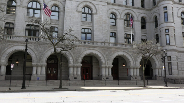 Two U.S. flags on flagpoles wave over a balustrade above a series of arches framing multiple doorways and windows on the ground floor of a multi-story Richardsonian Romanesque-style masonry building with turrets and arched windows, with leafless trees standing in the sidewalk next to a street. Two U.S. flags on flagpoles wave over a balustrade above a series of arches framing multiple doorways and windows on the ground floor of a multi-story Richardsonian Romanesque-style masonry building with turrets and arched windows, with leafless trees standing in the sidewalk next to a street.