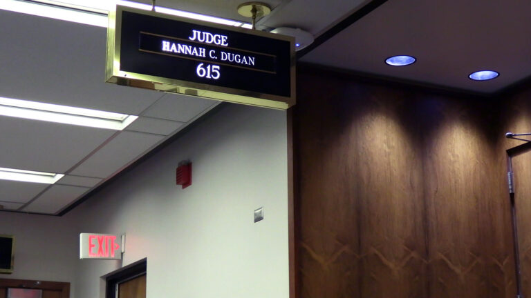 A ceiling-mounted brass enclosed letter sign with the words Judge Hannah C. Dugan and the number 615 is overhead in a hallway with fluorescent lighting outside the wood-paneled doors to a room, with an illuminated EXIT sign above another door in the background.
