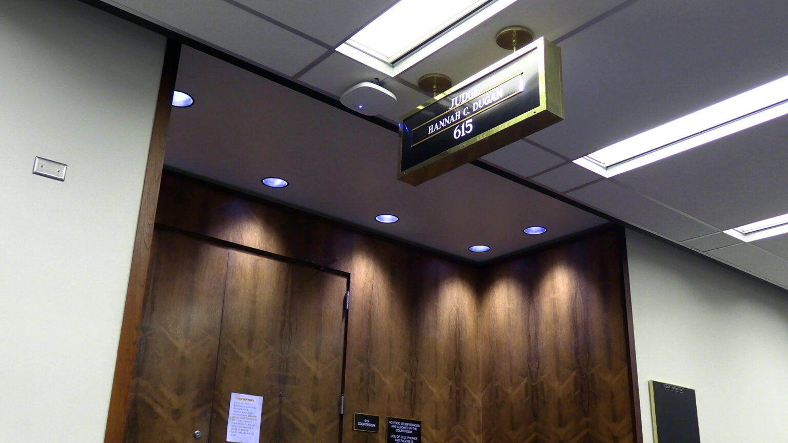 A ceiling-mounted brass enclosed letter sign with the words Judge Hannah C. Dugan and the number 615 is overhead in a hallway with fluorescent lighting outside the wood-paneled entrance to a room, with other signs affixed on and next to the door.
