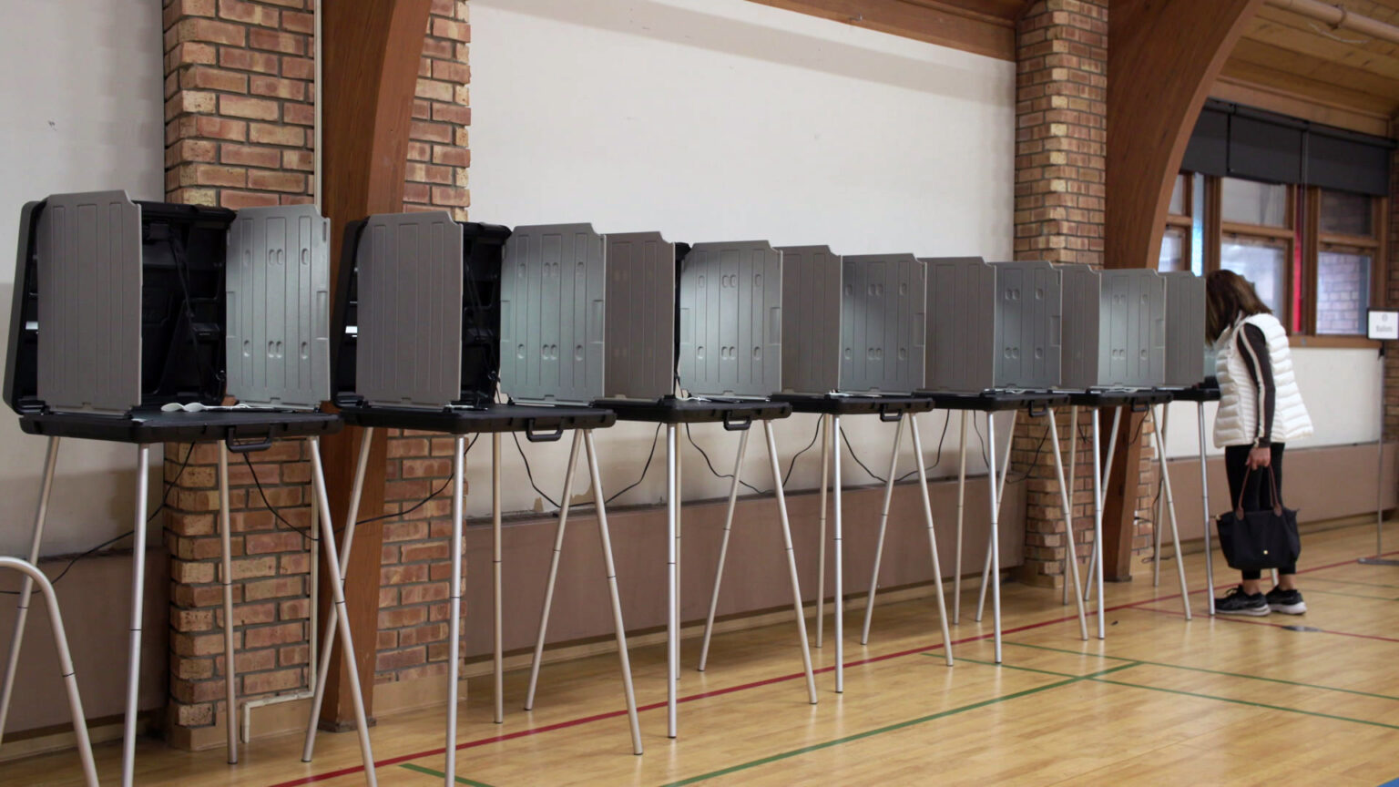 A voter holding the straps of a tote bag in their left hand stands in front of a temporary voting booth with plastic privacy walls and metal legs, located at the end of a row of similar booths in a gymnasium with a wood floor, brick columns and a wood-paneled vaulted ceiling.