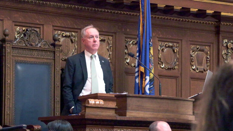Robin Vos stands and faces a microphone and gavel on a wood podium, with a high-backed leather and wood chair and a Wisconsin flag behind him, in a room with wood paneling featuring carved relief crests, with the out-of-focus heads of other people in the foreground. Robin Vos stands and faces a microphone and gavel on a wood podium, with a high-backed leather and wood chair and a Wisconsin flag behind him, in a room with wood paneling featuring carved relief crests, with the out-of-focus heads of other people in the foreground.