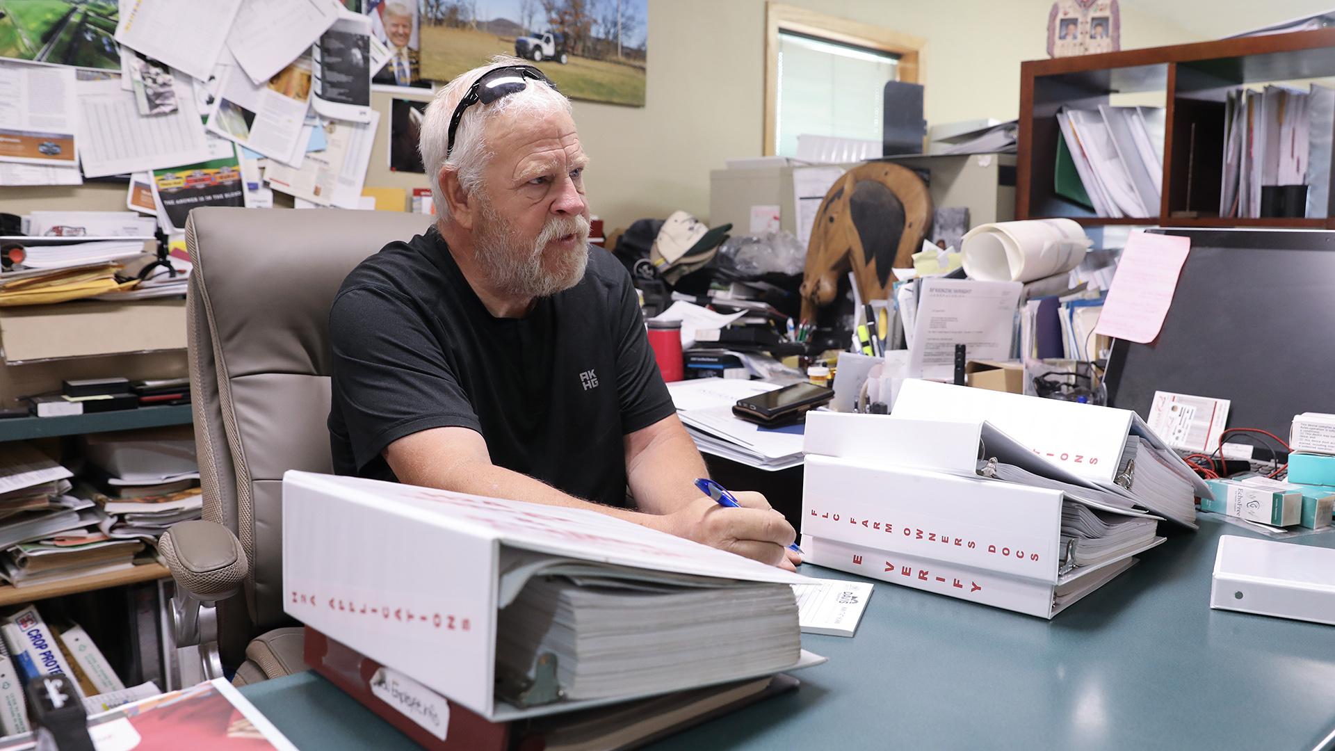 Mike Bushman sits in a high-backed leather chair and holds a pen in his right hand over a notepad on a desk in between stacks of three ring binders, with a computer monitor and stacks of other items on the desk's surface to his left, in a room with shelves filled with binders, papers and other items in the background.