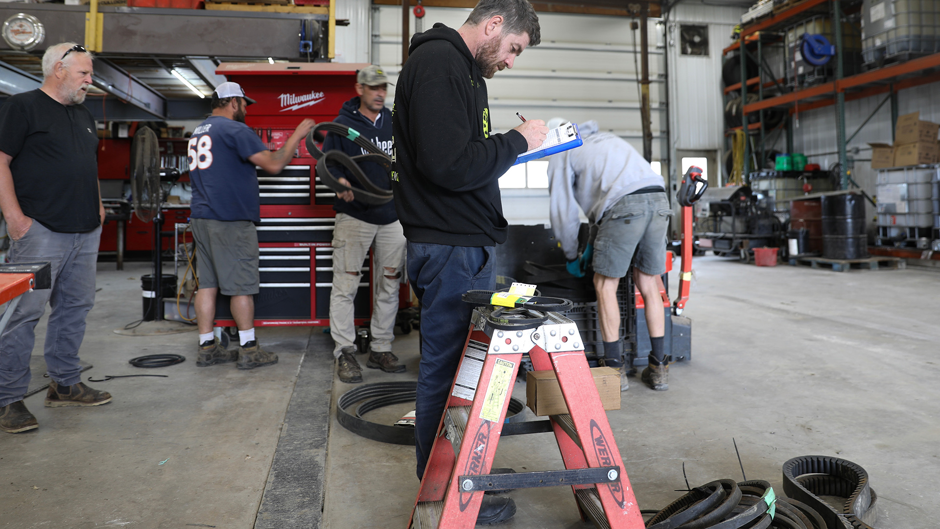 Monty Lilford stands and holds a pen in his right hand and writes on a sheet of paper connected to a clipboard, with four other people standing behind him and a short stepladder in front of him inside a high-ceilinged room with a concrete floor, tool cabinet, metal shelves filled with stacks of different containers and a metal garage door.