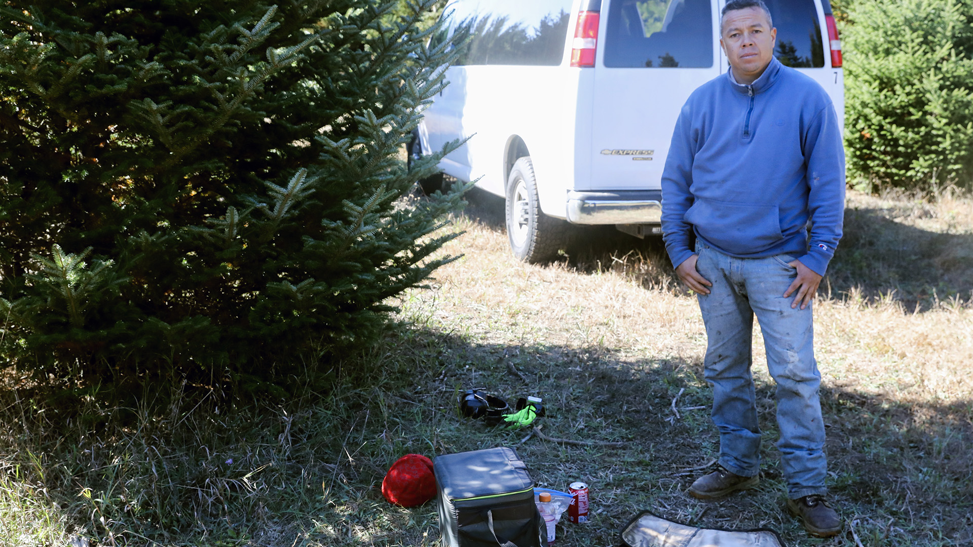 Roy Fernando Gonzalez Ramirez poses for a portrait while standing next to a coniferous tree, with a cooler, beverage containers, gloves and other items on the ground in front of him, and a parked van and more trees behind him in the background.