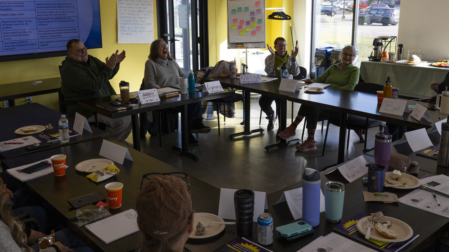 A group of people sit on the outside of four tables with metal legs and laminate surfaces arranged in a square, with plates of food, beverage containers, phones, papers and paper name plats on their surfaces, in a room with a large monitor mounted on a wall next to a metal and glass door, with a table with food and beverages on its surface in front of a window on an adjacent wall.