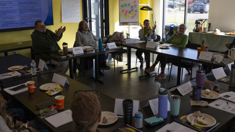 A group of people sit on the outside of four tables with metal legs and laminate surfaces arranged in a square, with plates of food, beverage containers, phones, papers and paper name plats on their surfaces, in a room with a large monitor mounted on a wall next to a metal and glass door, with a table with food and beverages on its surface in front of a window on an adjacent wall.