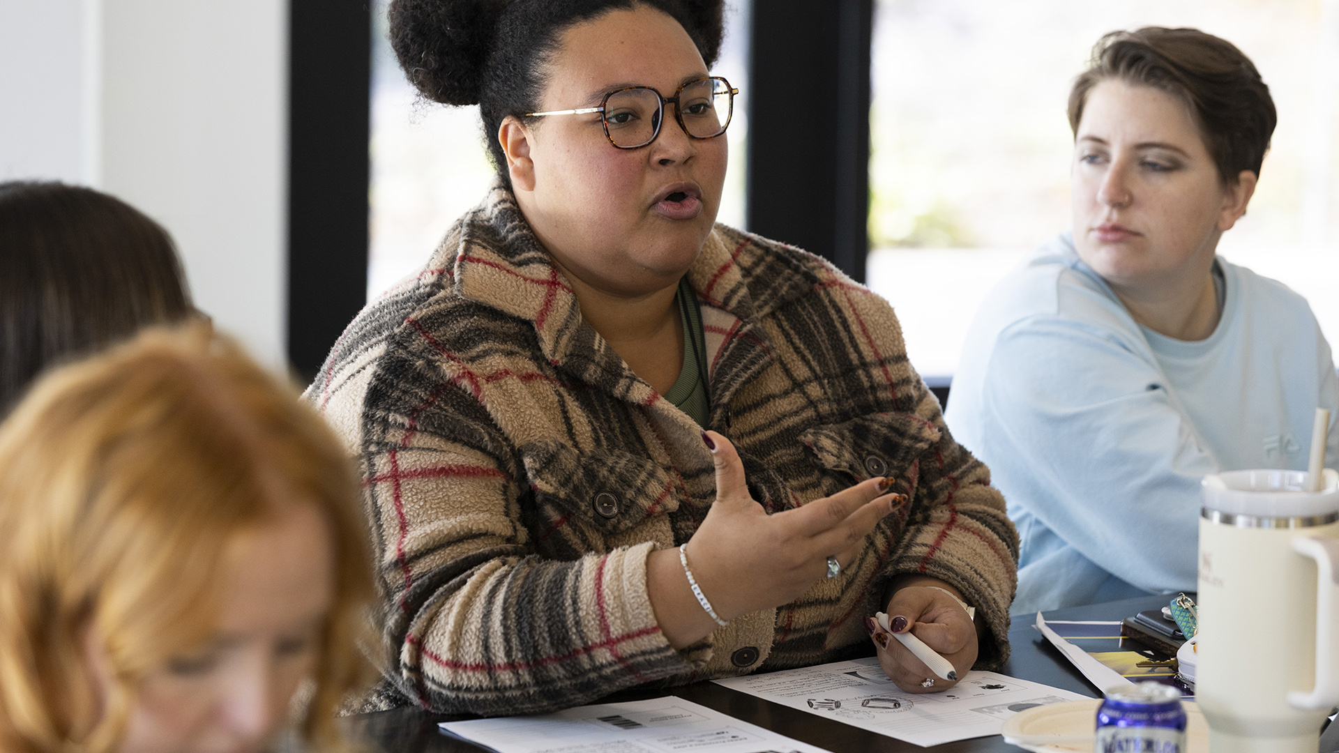 Caprice Swanks gestures with her right hand and holds a pen in her left hand while speaking and sitting at a table with papers, beverage containers and other items on its surface, with other out-of-focus people seated on either side and in front of her, in a room with windows in the background,