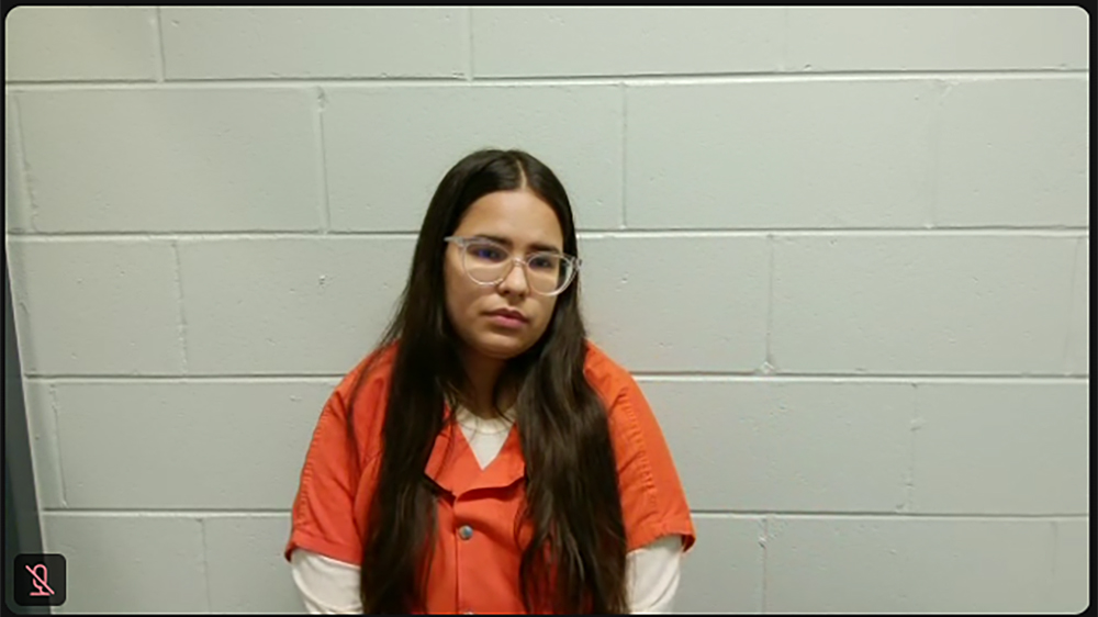 A screenshot of a video shows Dailin Pacheco-Acosta sitting in front of a painted concrete block wall, with an icon of a microphone with a line through it in the lower left corner.