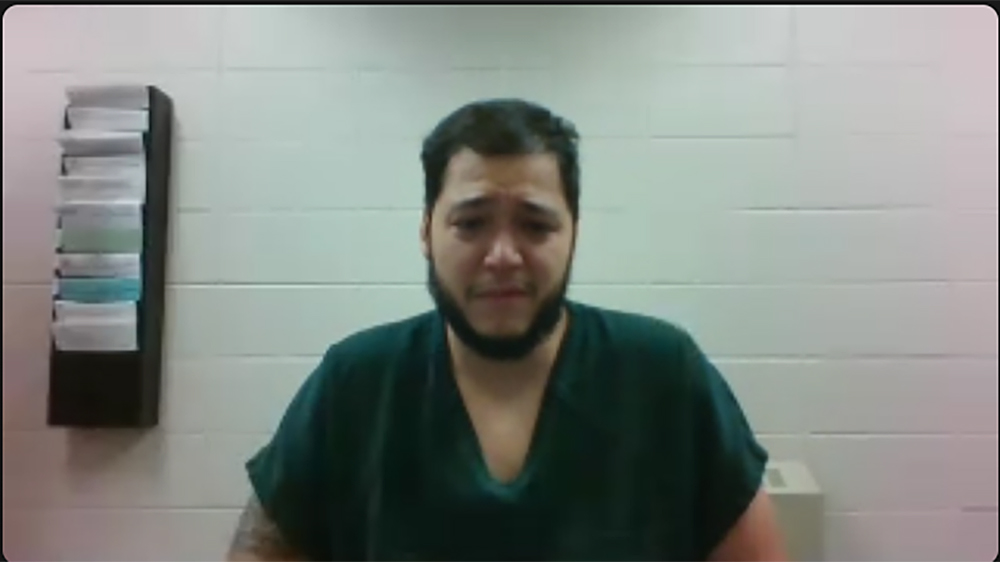 A screenshot of a video shows Dailin Pacheco-Acosta sitting in front of a painted concrete block wall with wall-mounted plastic paper organizer filled with different forms on one side.