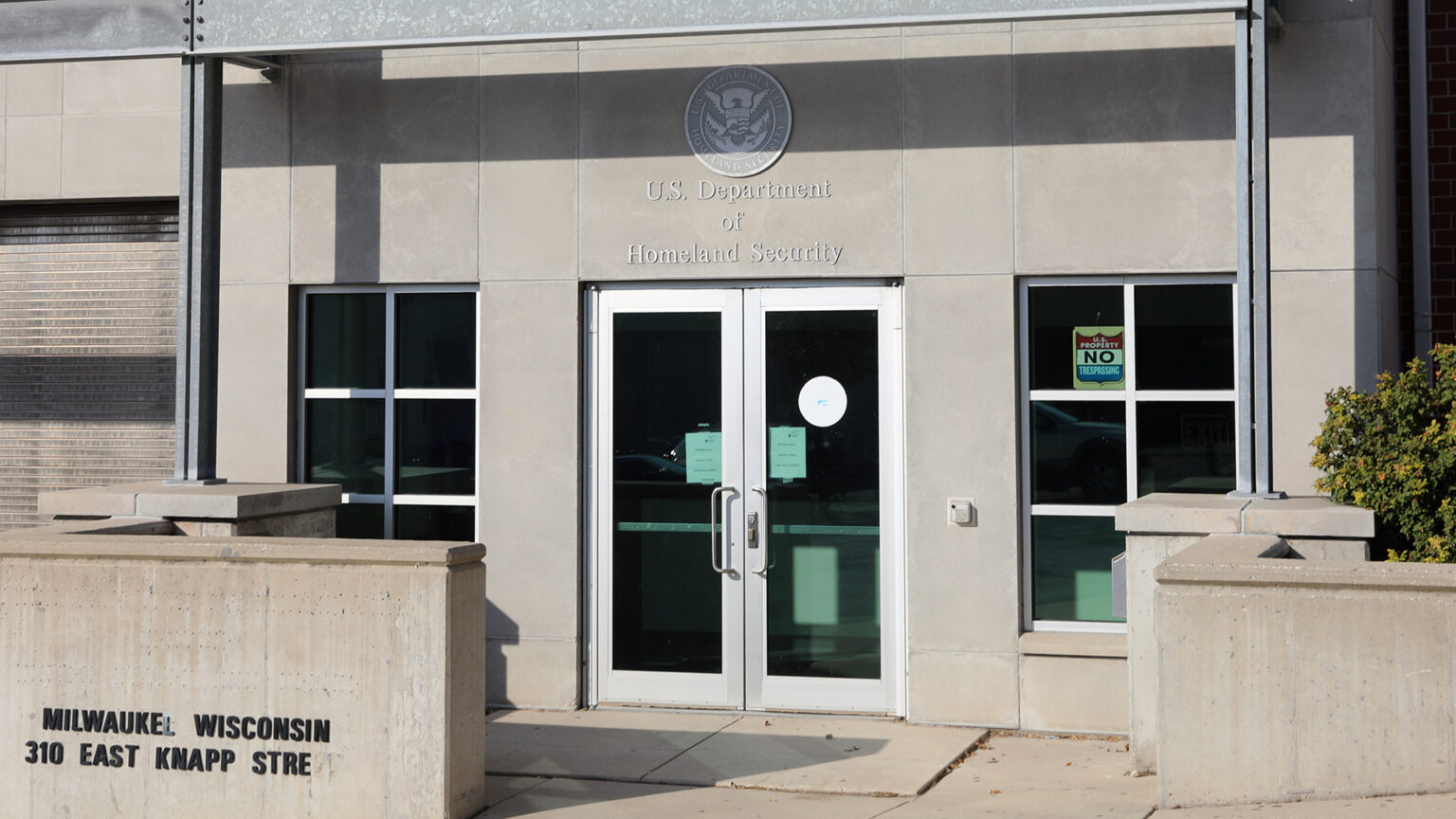 A letter sign on reading Milwauke Wisconsin and 310 East Knapp Stre is affixed to a concrete wall that's situated along a sidewalk and in front of a multistory building with a stone exterior and windows on either side of double glass doors below a letter sign reading U.S. Department of Homeland Security.