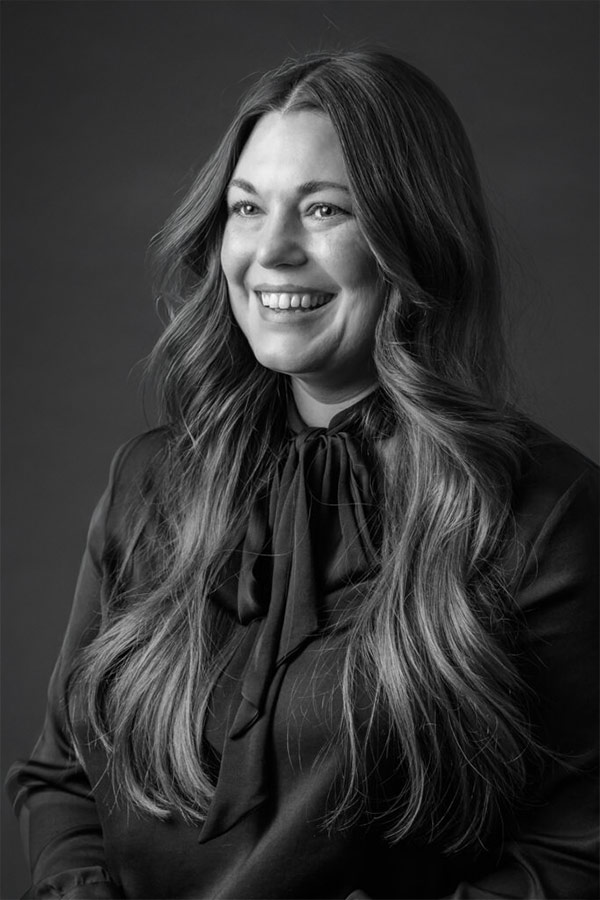 Adrienne Palm Smiling woman with long wavy hair wearing a dark blouse with a bow tie collar, posed against a dark studio background.