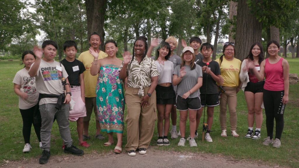 A group of people stands outdoors on grass, smiling and waving. Trees in the background suggest a park setting, creating a joyful, relaxed atmosphere.