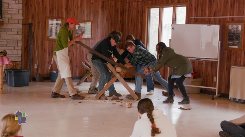 A group of six people enthusiastically saw a log with a large handsaw inside a rustic wooden room. Onlookers sit on the floor, watching intently.