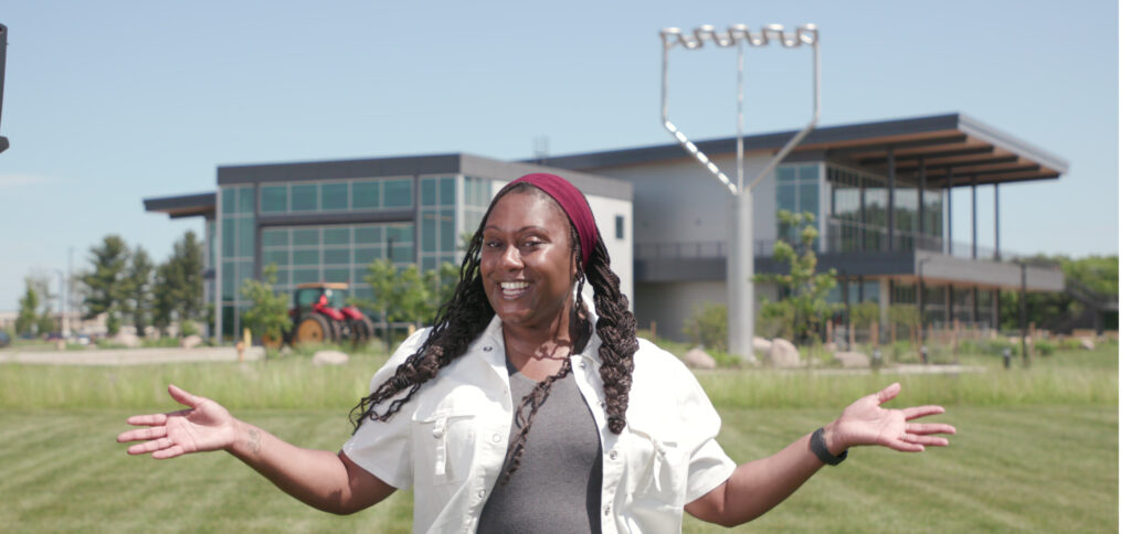 Smiling woman stands with arms outstretched in a grassy area. Modern building with large windows in the background. Bright, sunny day.
