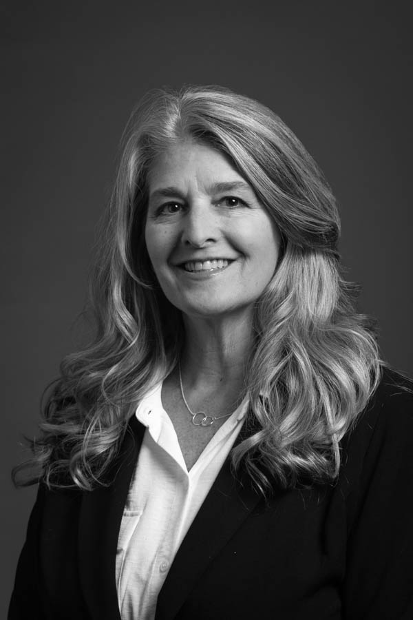 Natalie Glumm Smiling woman with long wavy hair wearing a blazer over a button-up shirt and a delicate necklace, posed against a dark background.