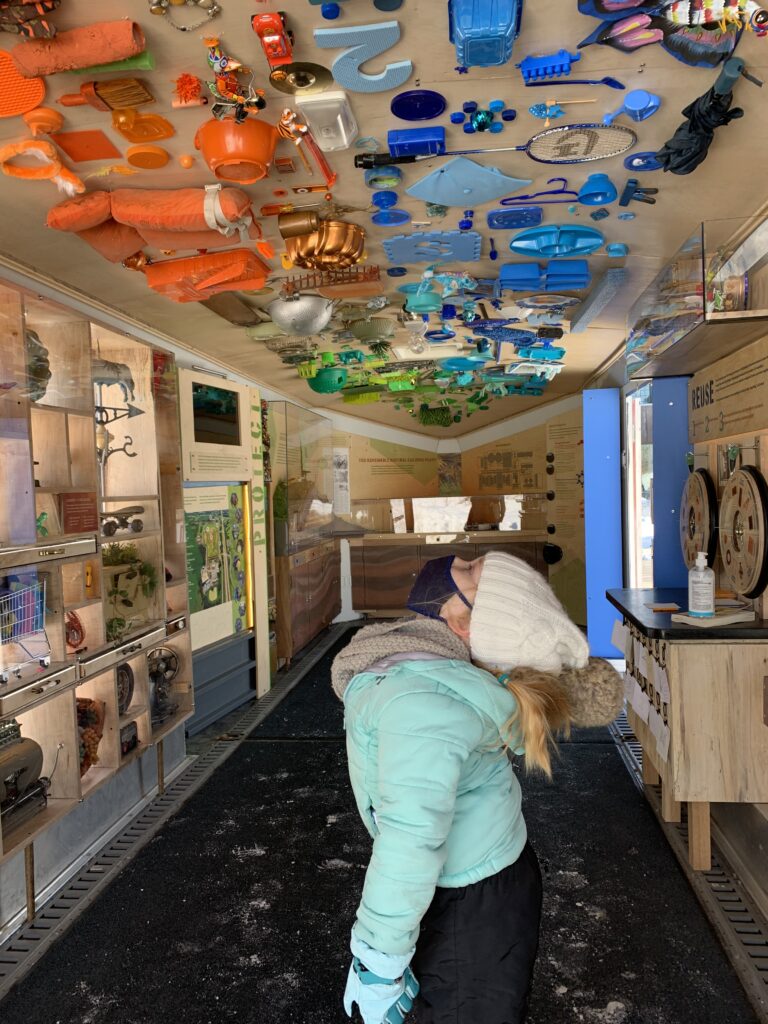 A child in a winter coat and hat gazes upward to the ceiling of the Dane County Trash Lab exhibit filled with colorful recycled objects, including toys and containers, mounted on the ceiling. The scene is educational and whimsical.