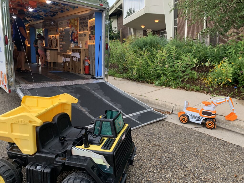 Toy trucks near an open trailer of the Dane County Trash Lab on a street, with people browsing inside. Lush greenery lines the sidewalk, creating a lively, casual scene.