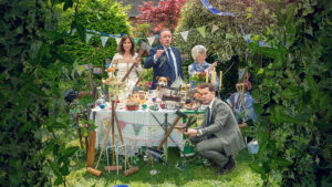 A whimsical garden tea party frozen as a crime scene: five adults pose among cakes, teacups and tools, with police tape strung through greenery, a small dog sitting on the table, and one man crouching with a hammer amid lush foliage.