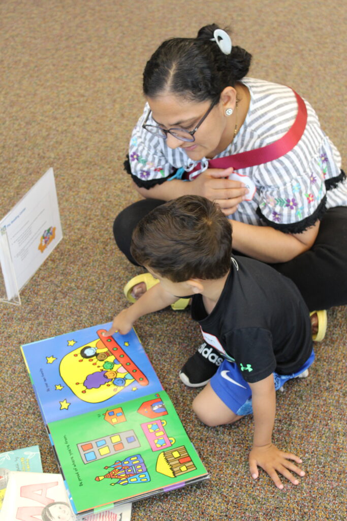 A caregiver and young child sit on a floor reading books together