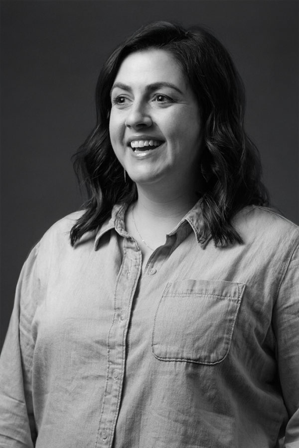Nikki Hessel Smiling woman with shoulder-length wavy hair wearing a button-up shirt with a front pocket, posed against a dark studio background.