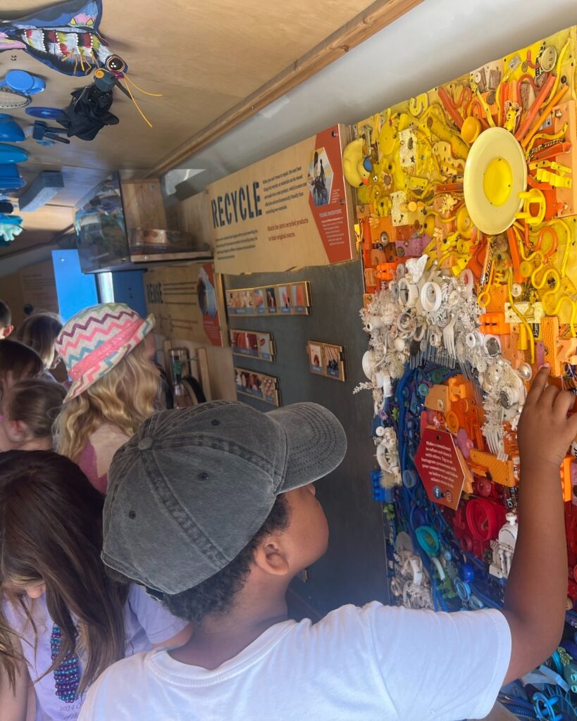Children explore the Dane County Trash Lab at the MSCR Camp Compass, a vibrant recycling art exhibit filled with colorful items like bottle caps. The mood is educational and engaging, with focused expressions. 