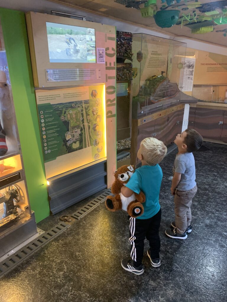 Two children are captivated by a nature exhibit in a museum in the Dane County Trash Lab at the 2025 Garden & Green Living Expo. One holds a stuffed animal while they both gaze at an illuminated screen displaying wildlife.