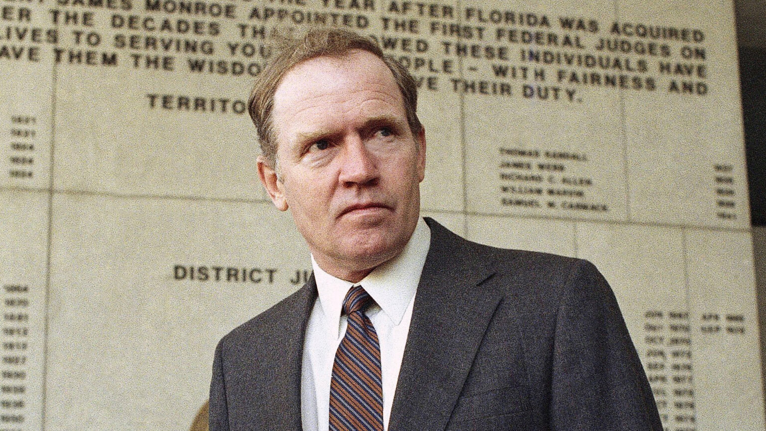 Eugene Hasenfus stands in front of a stone wall with multiple lines of letter sign text with names and dates.