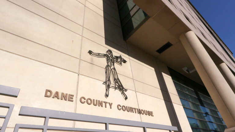 A sign reading County Courthouse and a stylized flat, wire sculpture of Lady Justice are mounted on the masonry wall of a Brutalist-style building with a row of pillars in front of glass windows. A sign reading County Courthouse and a stylized flat, wire sculpture of Lady Justice are mounted on the masonry wall of a Brutalist-style building with a row of pillars in front of glass windows.