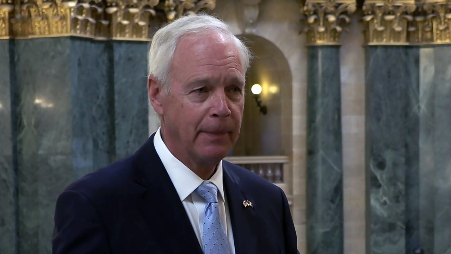 Ron Johnson stands and speaks inside a building with out-of-focus marble masonry and pillars with Composite order capitals in the background. Ron Johnson stands and speaks inside a building with out-of-focus marble masonry and pillars with Composite order capitals in the background.