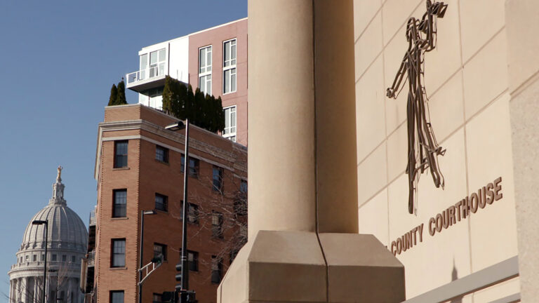 A sign reading County Courthouse and a stylized flat, wire sculpture of Lady Justice are mounted on the masonry wall of a Brutalist-style building, with multi-story brick and metal-sided buildings across an intersection and on either side of a street, with the Wisconsin State Capitol in the background under a clear sky. A sign reading County Courthouse and a stylized flat, wire sculpture of Lady Justice are mounted on the masonry wall of a Brutalist-style building, with multi-story brick and metal-sided buildings across an intersection and on either side of a street, with the Wisconsin State Capitol in the background under a clear sky.