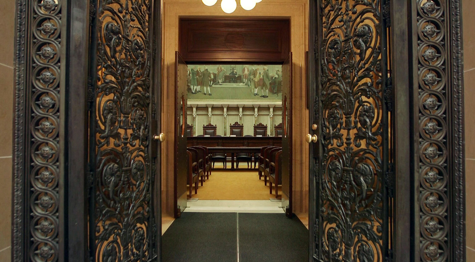 Metal filigree double doors with brass doorknobs and lock mechanisms open into an antechamber with an illuminated overhead light fixture, commercial floor mats and a set of wood double doors with oval glass windows opening into a room with a judicial bench with two sets of seats faced by a low table and multiple rows of wood and leather chairs in a room with marble masonry and a large painting.