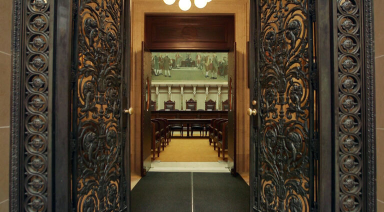 Metal filigree double doors with brass doorknobs and lock mechanisms open into an antechamber with an illuminated overhead light fixture, commercial floor mats and a set of wood double doors with oval glass windows opening into a room with a judicial bench with two sets of seats faced by a low table and multiple rows of wood and leather chairs in a room with marble masonry and a large painting.