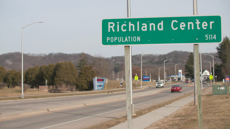 A city limit sign with the words Richland Center and Population 5114 is affixed to wood poles next to a sidewalk and divided highway with vehicles traveling in both directions, with trees, signs and a gas station in the background below low hills on the horizon. A city limit sign with the words Richland Center and Population 5114 is affixed to wood poles next to a sidewalk and divided highway with vehicles traveling in both directions, with trees, signs and a gas station in the background below low hills on the horizon.