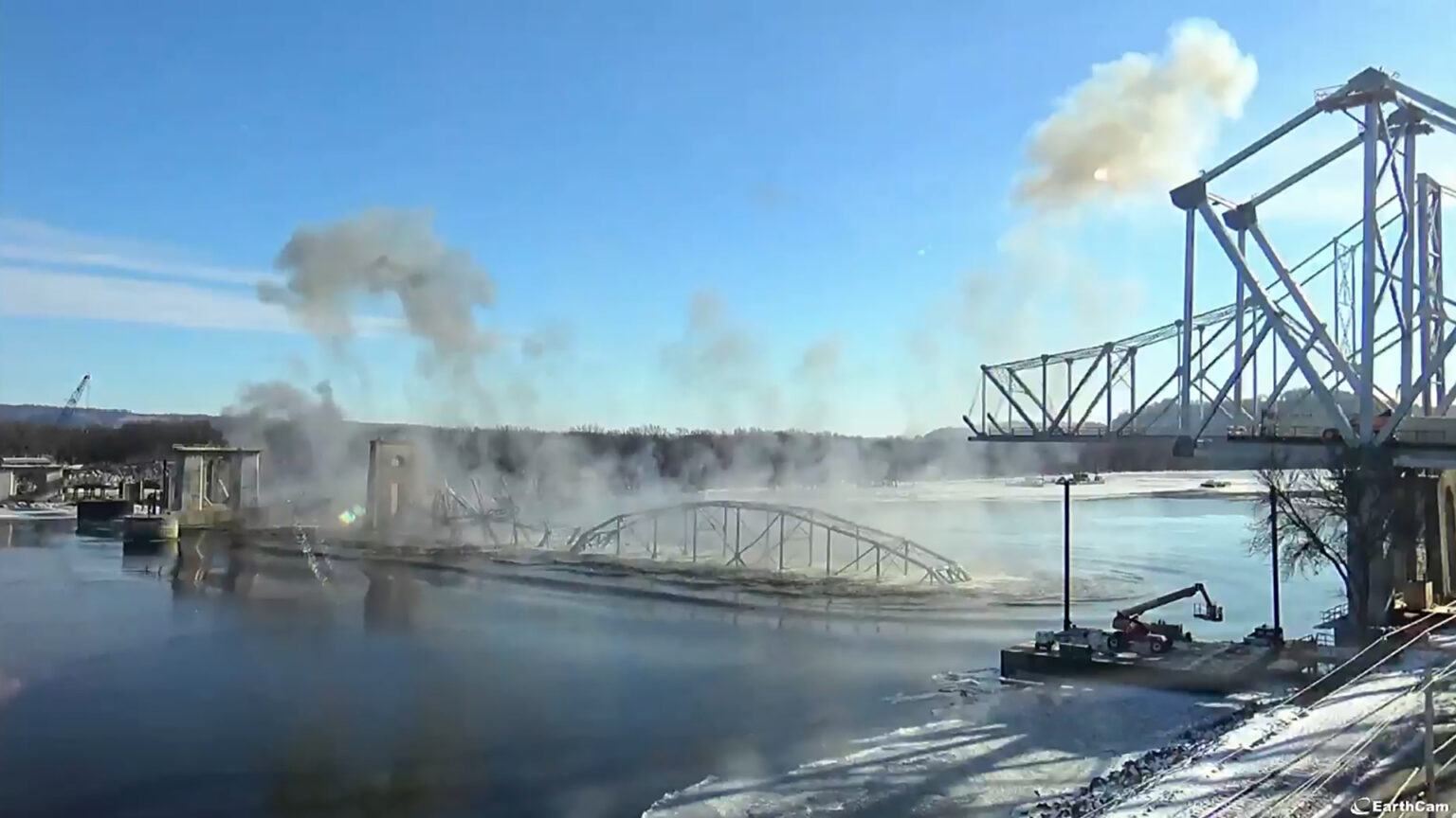 The center and one side span of riveted cantilever metal bridge land on the surface of a river with clouds of dust rising into the air and obscuring the sun, with another span to the side still standing along with multiple concrete support structures, with ice along the edges of the water and wooded areas on the far shore.