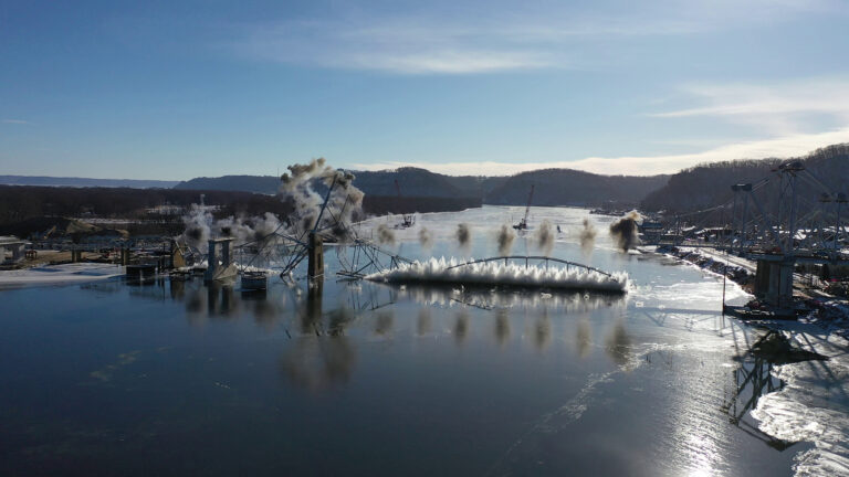 The center and one side span of riveted cantilever metal bridge splash on the surface of a river with clouds of dust rising into the air, with another span to the side still standing along with multiple concrete support structures, with ice downriver and along the edges of the shore and with wooded bluffs in the background.