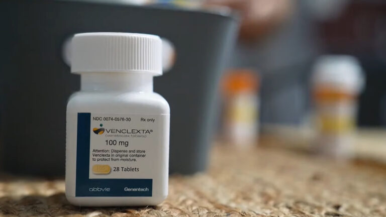 A plastic medication bottle with a label reading Venclexta®, 100 mg and 28 tablets sits on a woven fiber surface, with two out-of-focus medication vials in the background.
