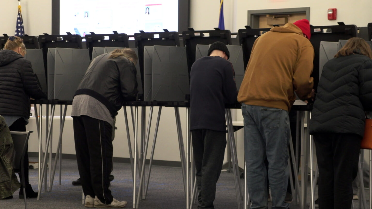 Five people stand and lean over while filling out ballots inside collapsible voting booths with metal legs and plastic tables and privacy walls, in a room with the U.S. and Wisconsin flags on either side of a monitor and next to a closed wood door with a hydraulic closer. Five people stand and lean over while filling out ballots inside collapsible voting booths with metal legs and plastic tables and privacy walls, in a room with the U.S. and Wisconsin flags on either side of a monitor and next to a closed wood door with a hydraulic closer.