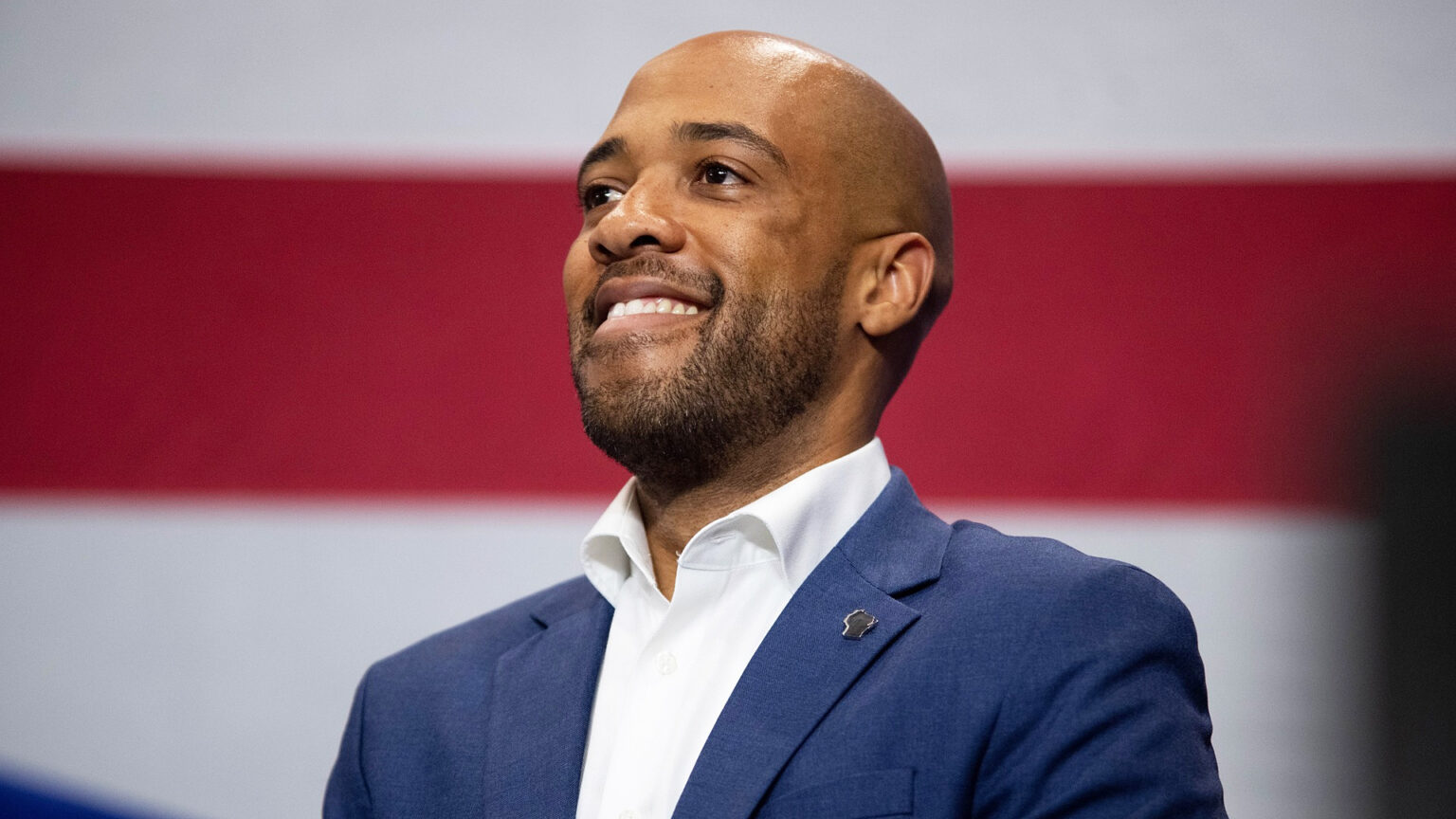 Mandela Barnes smiles while standing in front of the out-of-focus stripes of a U.S. flag.