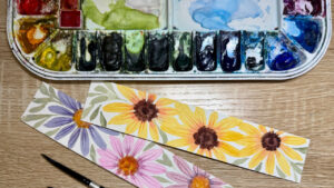 A set of watercolors used in an easel next to two bookmarks decorated with watercolored paintings of daisy flowers.