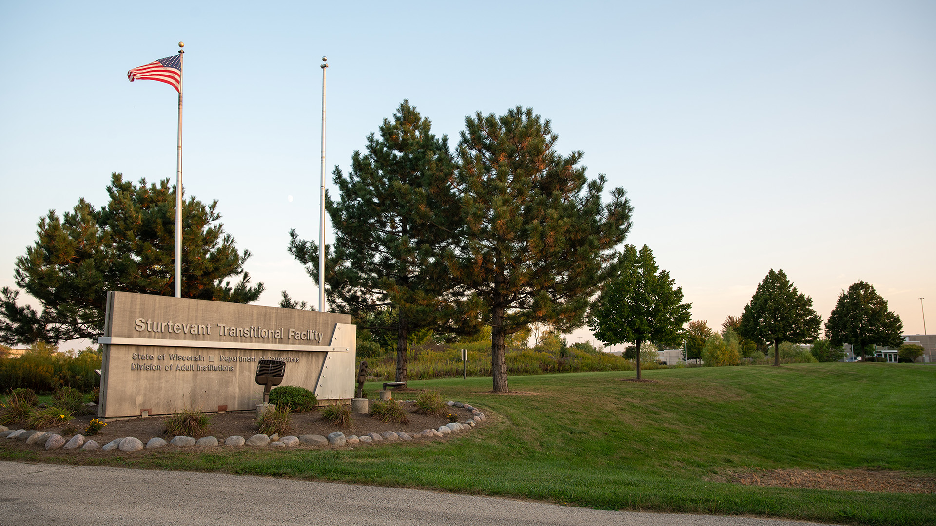 A U.S. flag on a flagpole waves in the wind next to an empty flagpole, with each standing behind a concrete wall with a letter sign on its surface reading "Sturtevant Transitional Facility," "State of Wisconsin Department of Corrections" and "Division of Adult Institutions" that is surrounded by landscape plants and a ring of small boulders, with multiple coniferous trees standing in a line on an adjacent lawn, with low buildings and more trees in the background.