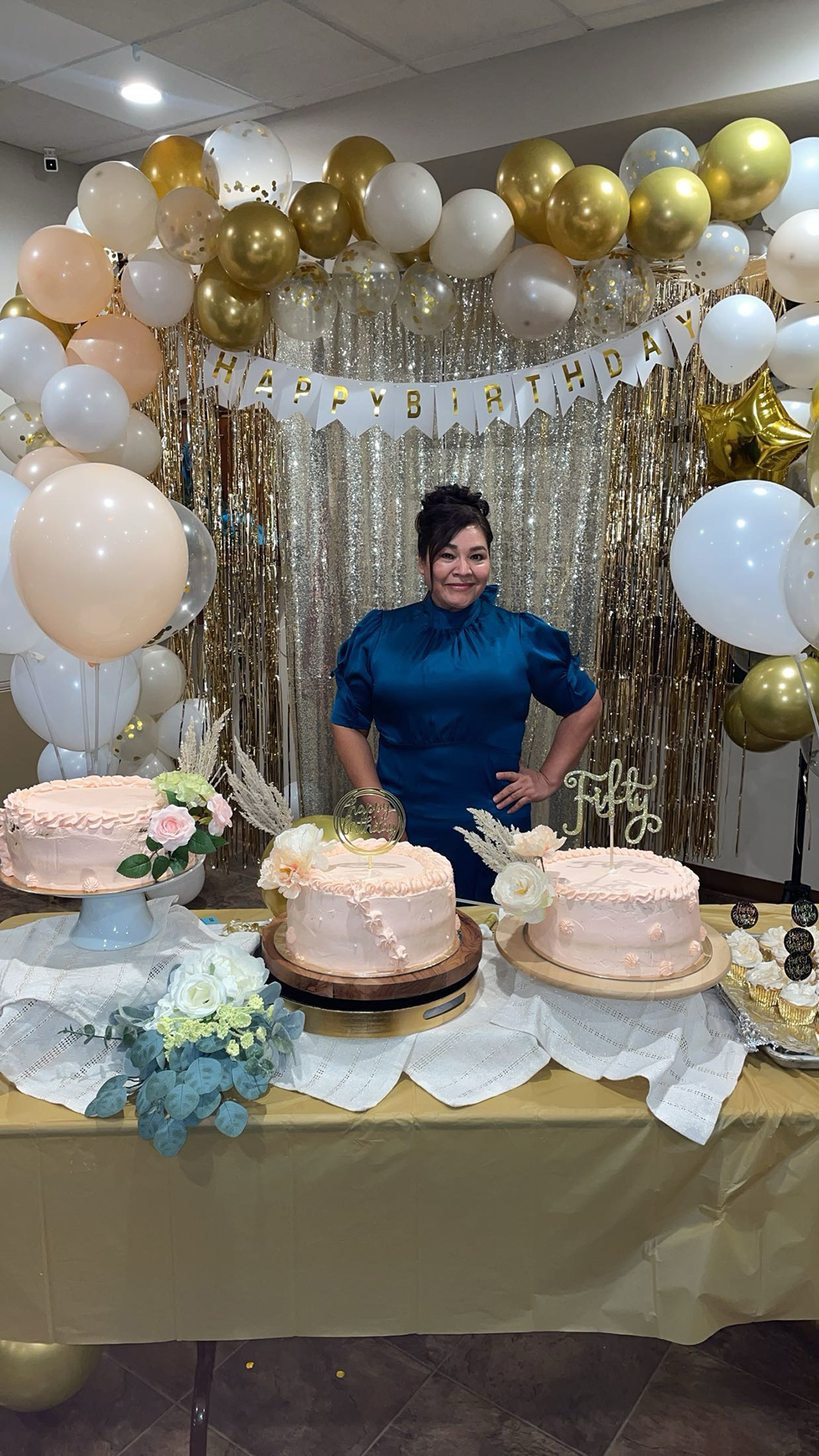 Elvira Benitez poses for a photo while standing behind a folding table with three round cakes on stands and a tray of cupcakes on its surface, with an arch of balloons, a party decoration banner with the words "Happy Birthday" and a tinsel curtain behind her in a room with a tile floor and drop-panel ceiling.