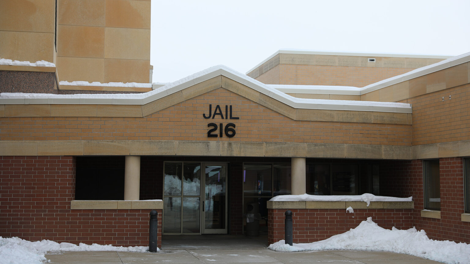 A letter sign with the word Jail and the numerals 216 are affixed to the surface of a brick wall above a sheltered patios with multiple glass doors to a multi-story brick and masonry building, with snow covering portions of different roof levels and areas of a paved patio.