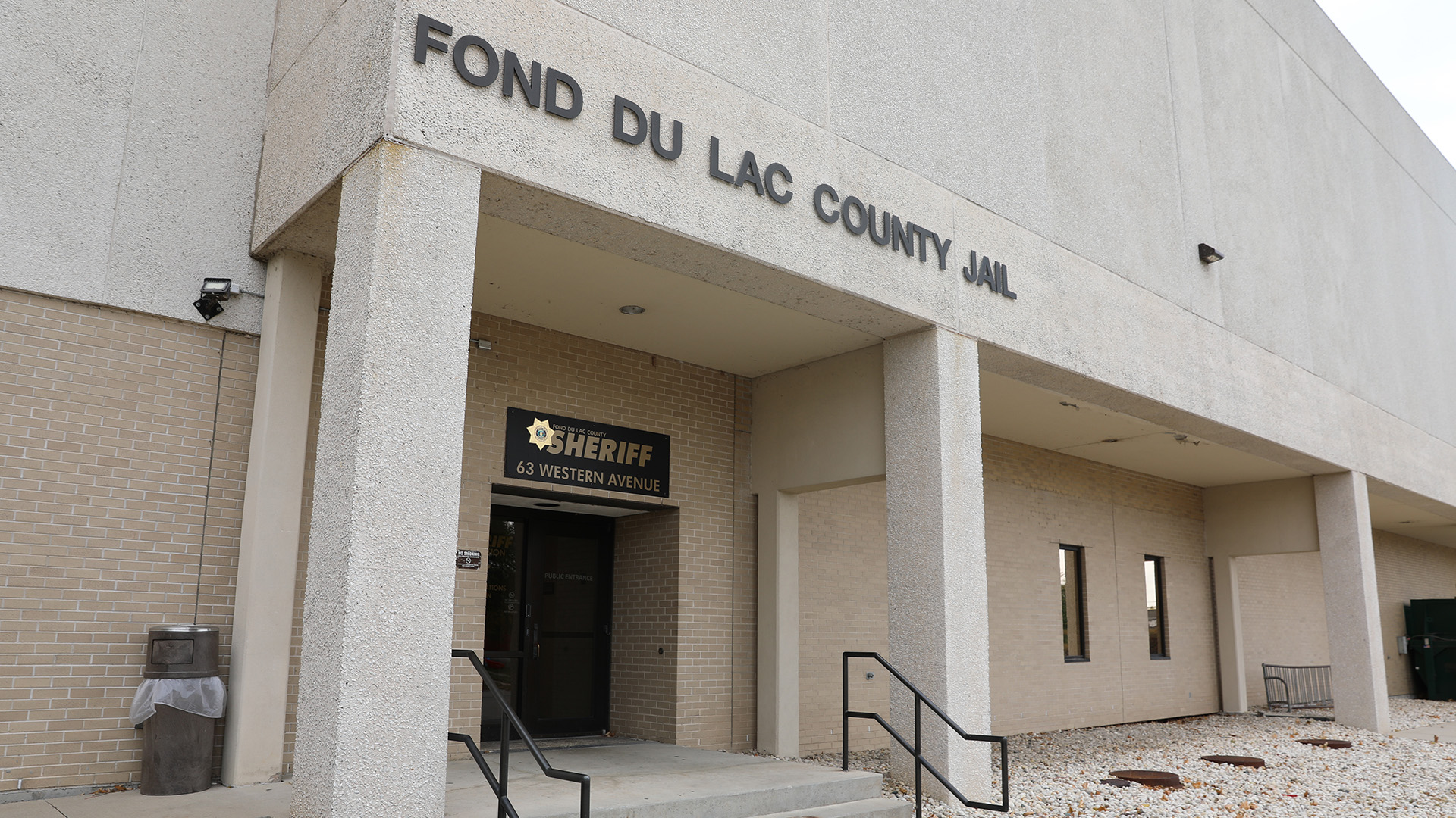 A letter sign reading "Fond du Lac County Jail" is affixed to the side of a multi-story brick and concrete building above an entrance with another sign showing a law enforcement badge and the words "Sheriff" and address "63 Western Avenue," with a short concrete staircase with metal railings leading to it next to a trash receptacle and front area covered with landscaping rock.